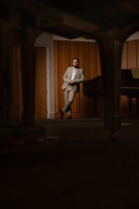 A man in a light blazer and tan trousers leans against a grand piano in a wood-panelled room, with warm lighting and the piano's blurred leg visible in the foreground.