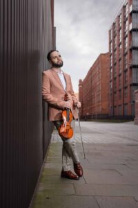 A man in a brown jacket leans against a dark wall on an empty city street, holding a violin and bow. He gazes thoughtfully upwards, with red-brick buildings and a cloudy sky in the background.