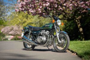A classic green Kawasaki z900 motorbike is parked on a paved path in a park, surrounded by blossoming trees with pink flowers and lush greenery.