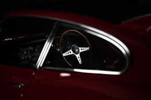 Close-up view of a classic red e type jaguar car’s interior, focusing on a wooden steering wheel, dashboard dials, and the side window outlined by chrome trim in low lighting.